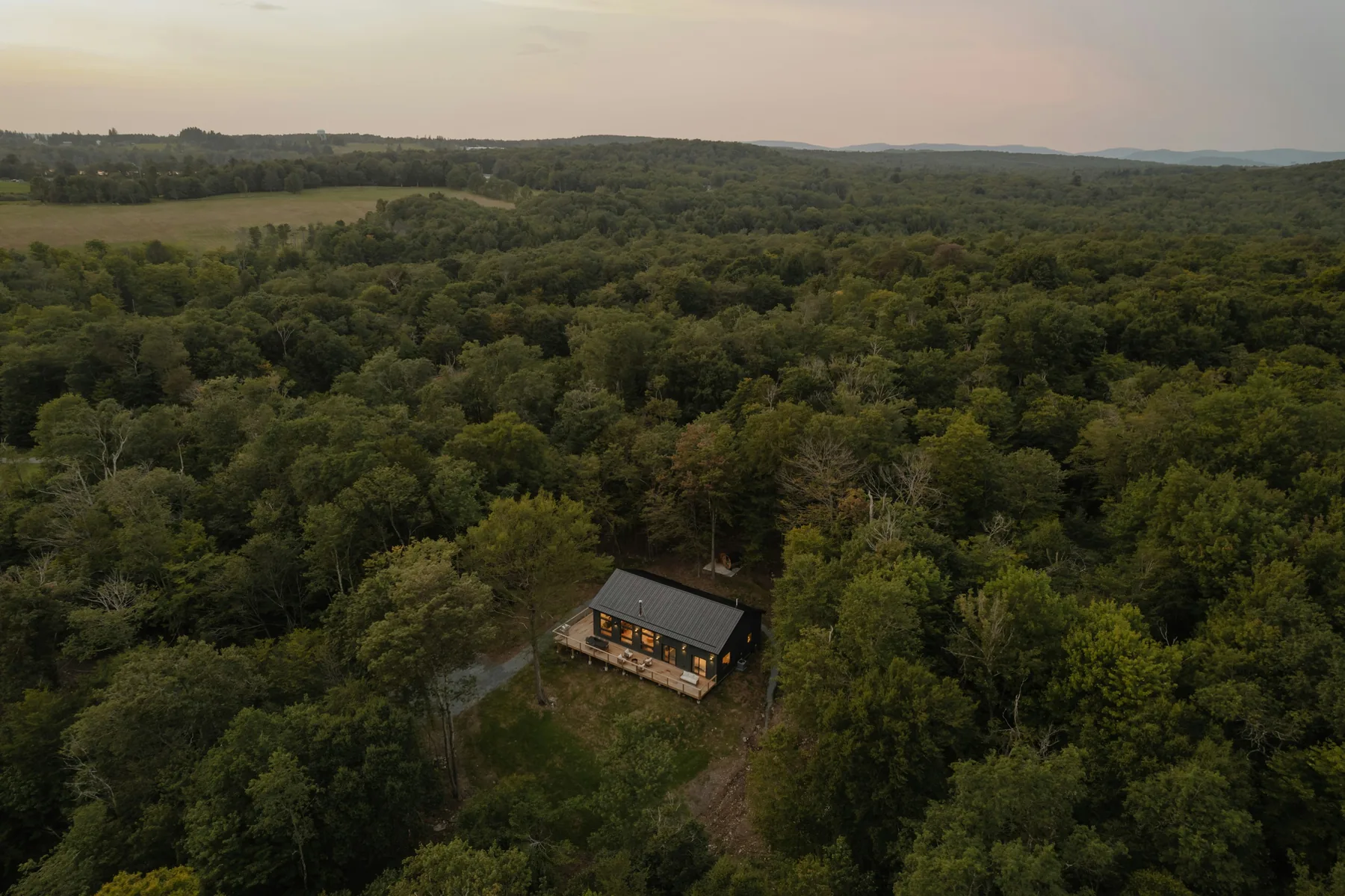 Luftaufnahme am Abend: ein einzelnes Holzhaus mit beleuchteten Fenstern, umgeben von Wald bis zum Horizont.